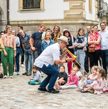 Posaunist spielt vor einer Reihe Kinder, die auf dem Boden sitzen. Um die Szene sind stehende Menschen versammelt.